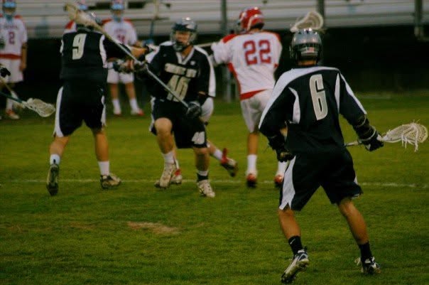 High school lacrosse game action, dark jerseys against a red and white team