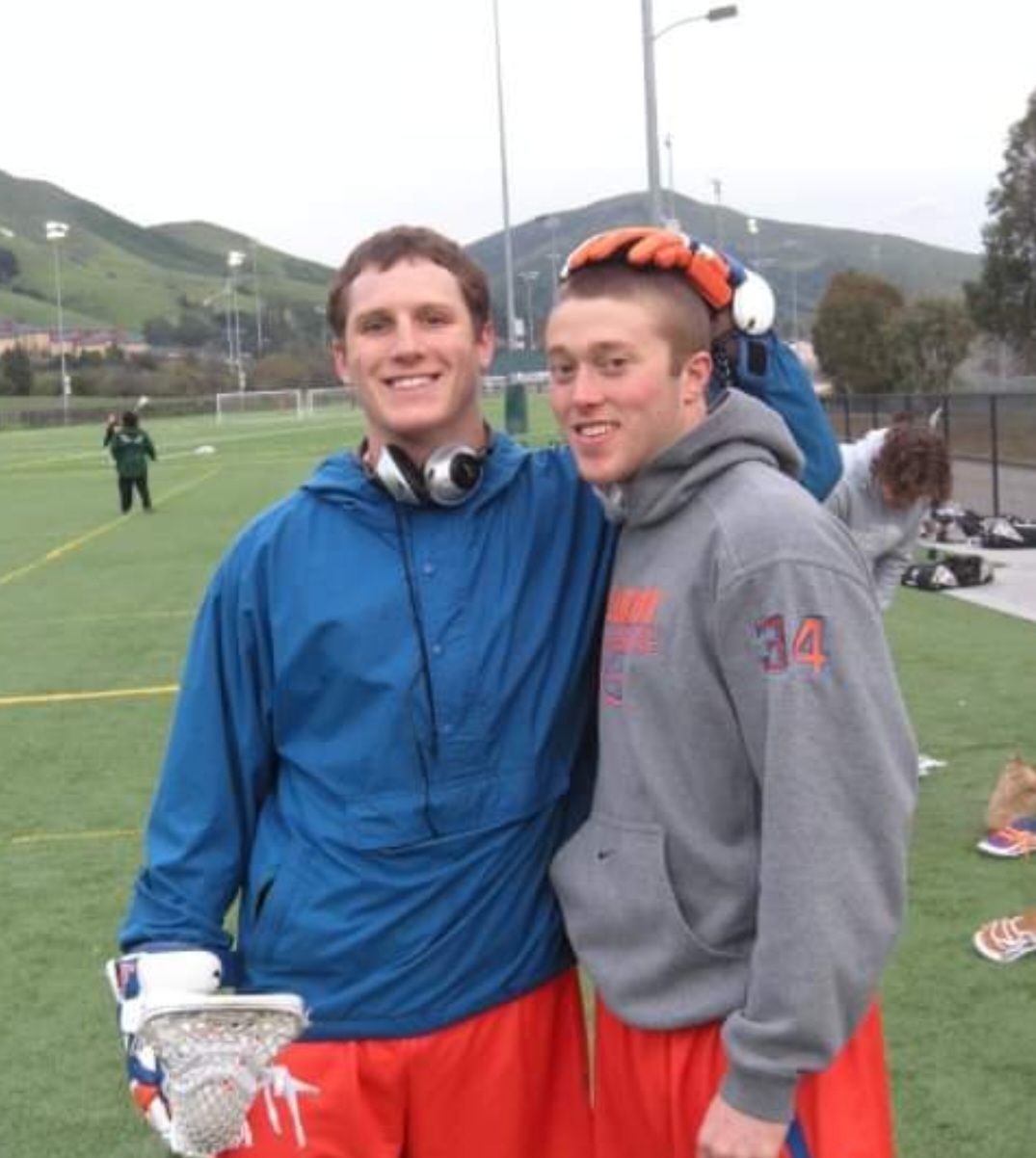 Peter Kimmel in a blue jacket holding a lacrosse stick, standing with a teammate during warmups
